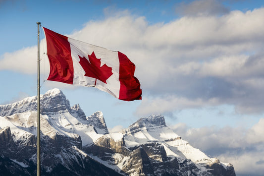 Canadian flag blowing in the wind on a flag pole with snow covered mountain range in the background with blue sky and clouds; Canmore, Alberta, Canada